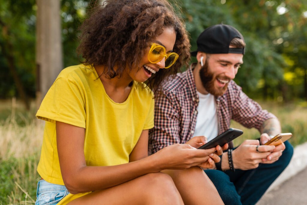happy young smiling friends sitting park using smartphones, man and woman having fun together, colorful summer hipster fashion style, communication wireless connecting devices